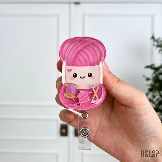 Close-up view of a knitting shaker badge reel featuring a smiling yarn ball with yarn spool, knitting needles, and knitted fabric charms.