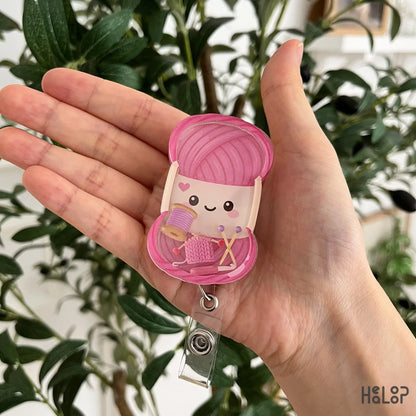 Cute knitting shaker badge reel featuring a smiling yarn ball with movable knitting-themed charms including yarn, knitting needles, and knitted fabric.