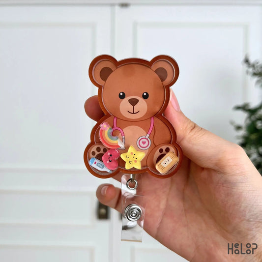 Detailed view of the Cute Teddy Bear Shaker Badge Reel featuring a smiling teddy bear with rainbow, heart, star, thermometer, and bandage shaker charms.