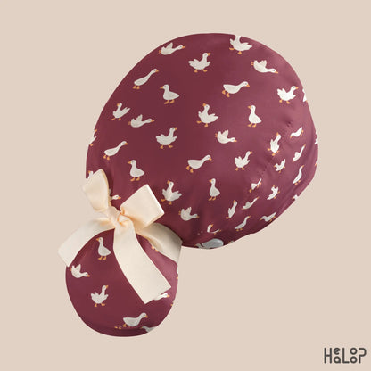 Angled rear view of the HEALOOP Goose Ponytail Scrub Cap with playful white geese on burgundy fabric, a cream-colored bow, and a rounded ponytail pouch.