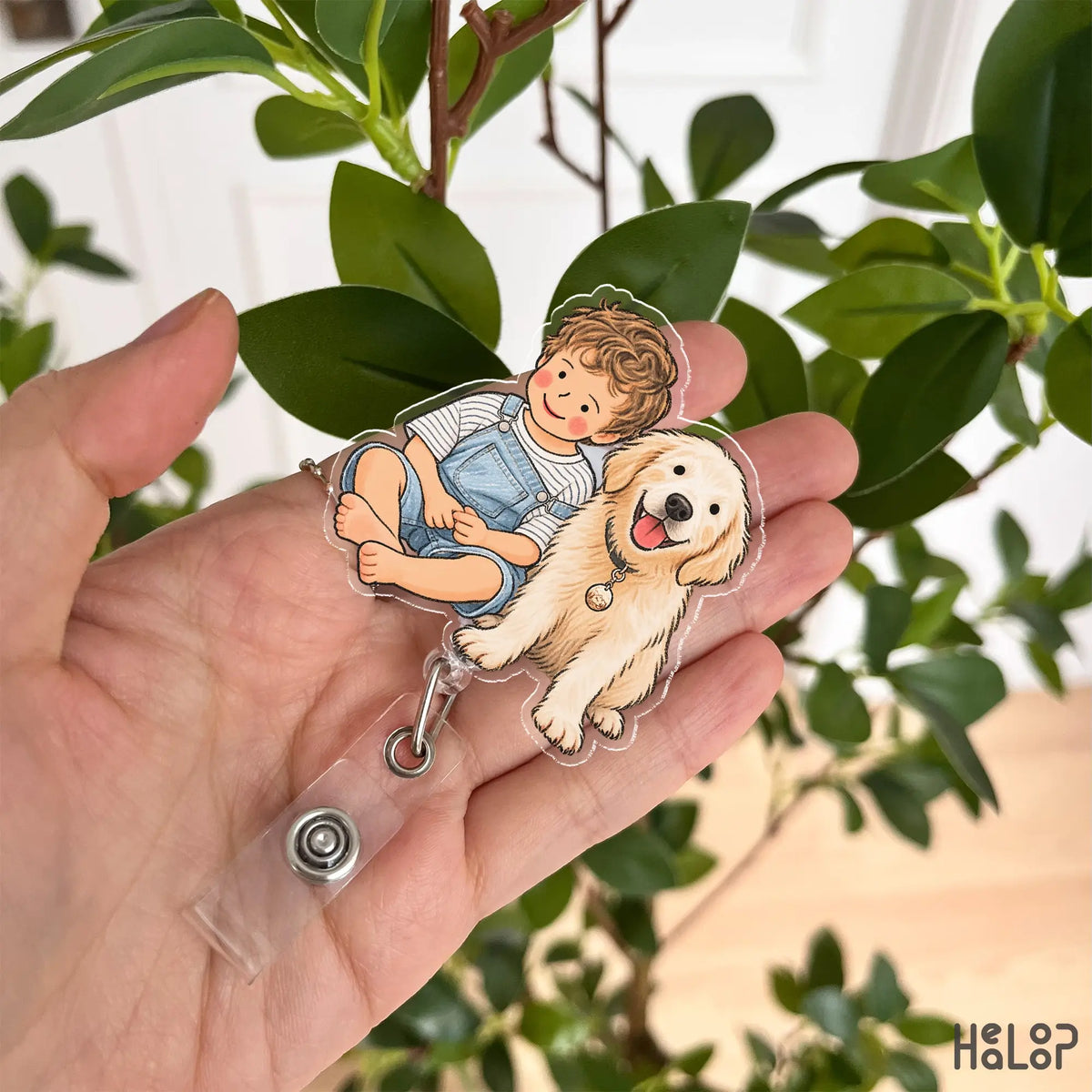 Close-up of a HEALOOP personalized portrait badge reel held in a hand, featuring a boy and golden retriever in a custom hand drawn design.