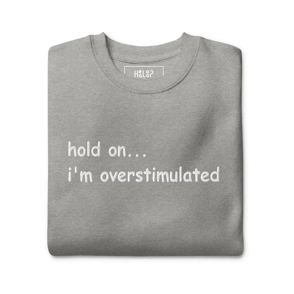 Folded 'Hold on... I’m overstimulated' sweatshirt in Carbon Grey, showcasing its cozy fabric and minimalist embroidered text design.