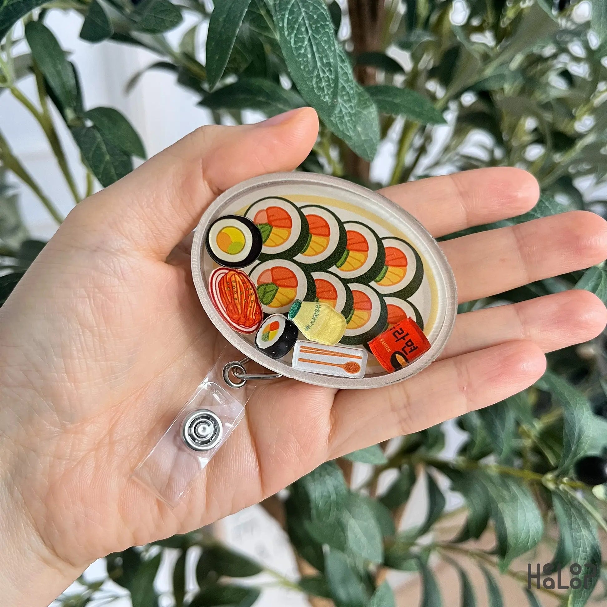 Handheld Korean kimbap shaker badge reel with vibrant K-food floating pieces.