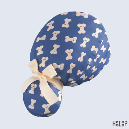 Orthopedic bone print ponytail scrub cap featuring playful cream-colored bone characters on blue fabric, shown from the side with a ribbon-tied ponytail pouch.
