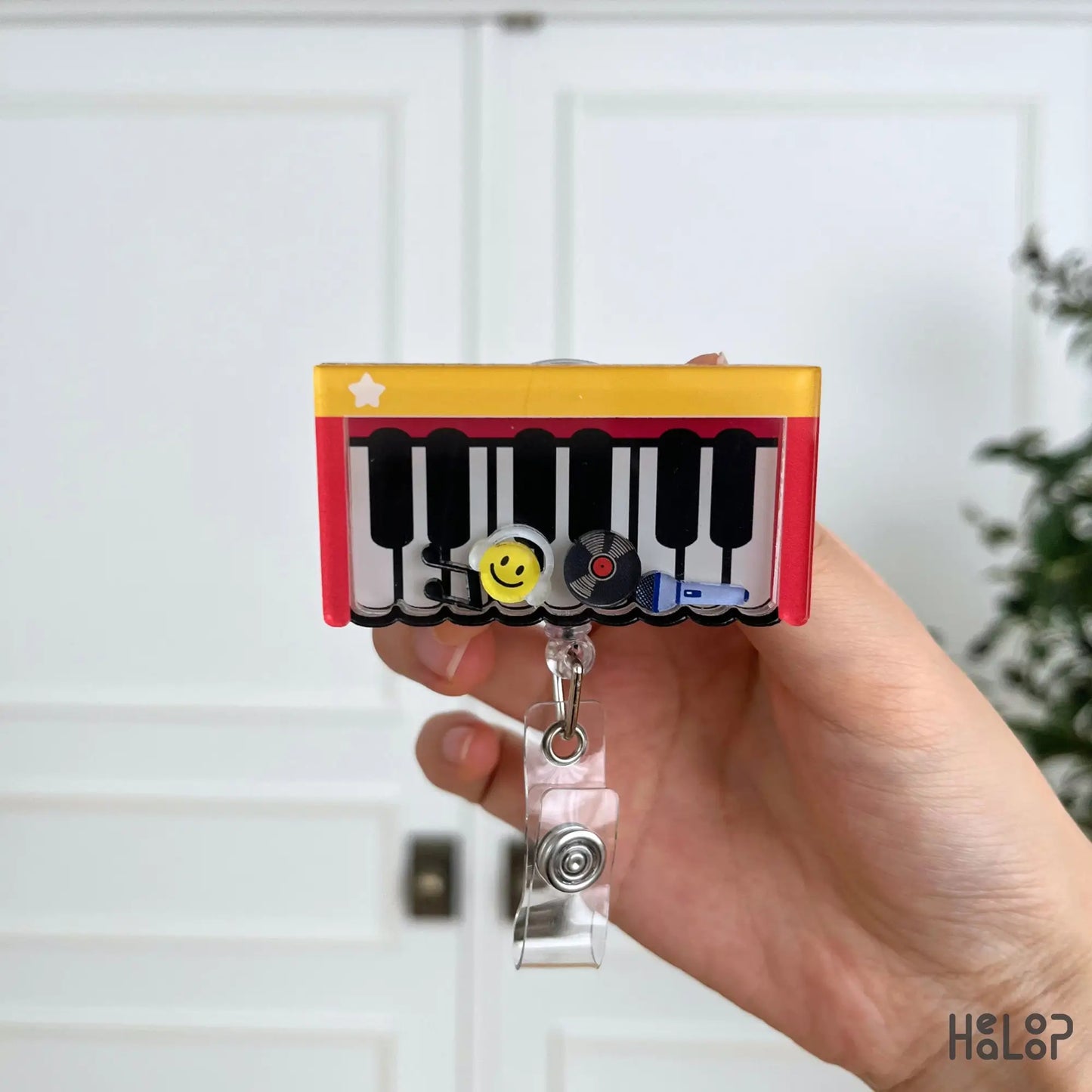 Cute Piano Shaker Badge Reel displayed in hand, featuring a piano keyboard with movable smiley face, vinyl record, music note, and microphone charms for music teachers and musicians.