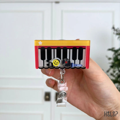 Cute Piano Shaker Badge Reel displayed in hand, featuring a piano keyboard with movable smiley face, vinyl record, music note, and microphone charms for music teachers and musicians.