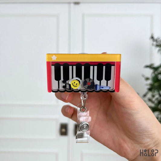 Cute Piano Shaker Badge Reel displayed in hand, featuring a piano keyboard with movable smiley face, vinyl record, music note, and microphone charms for music teachers and musicians.