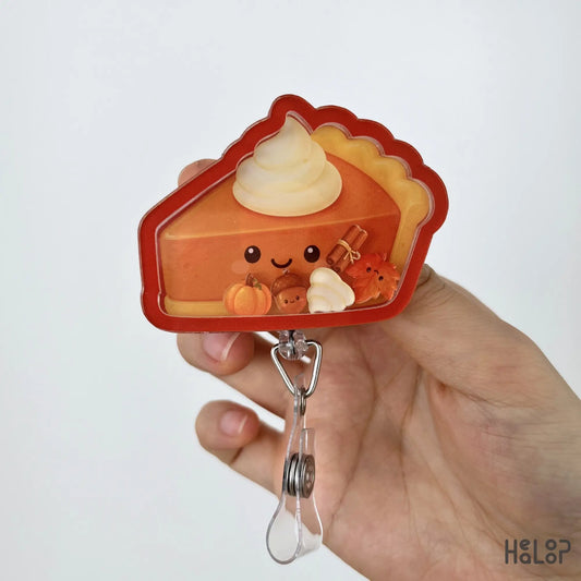 Close-up of the Pumpkin Pie Shaker Badge Reel with smiling pumpkin pie, whipped cream, pumpkin, acorn, cinnamon stick, and maple leaf shaker charms.