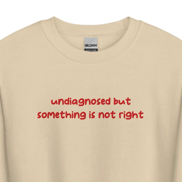 Sand-colored sweatshirt with 'undiagnosed but something is not right' embroidered in red, displayed flat for a detailed view.